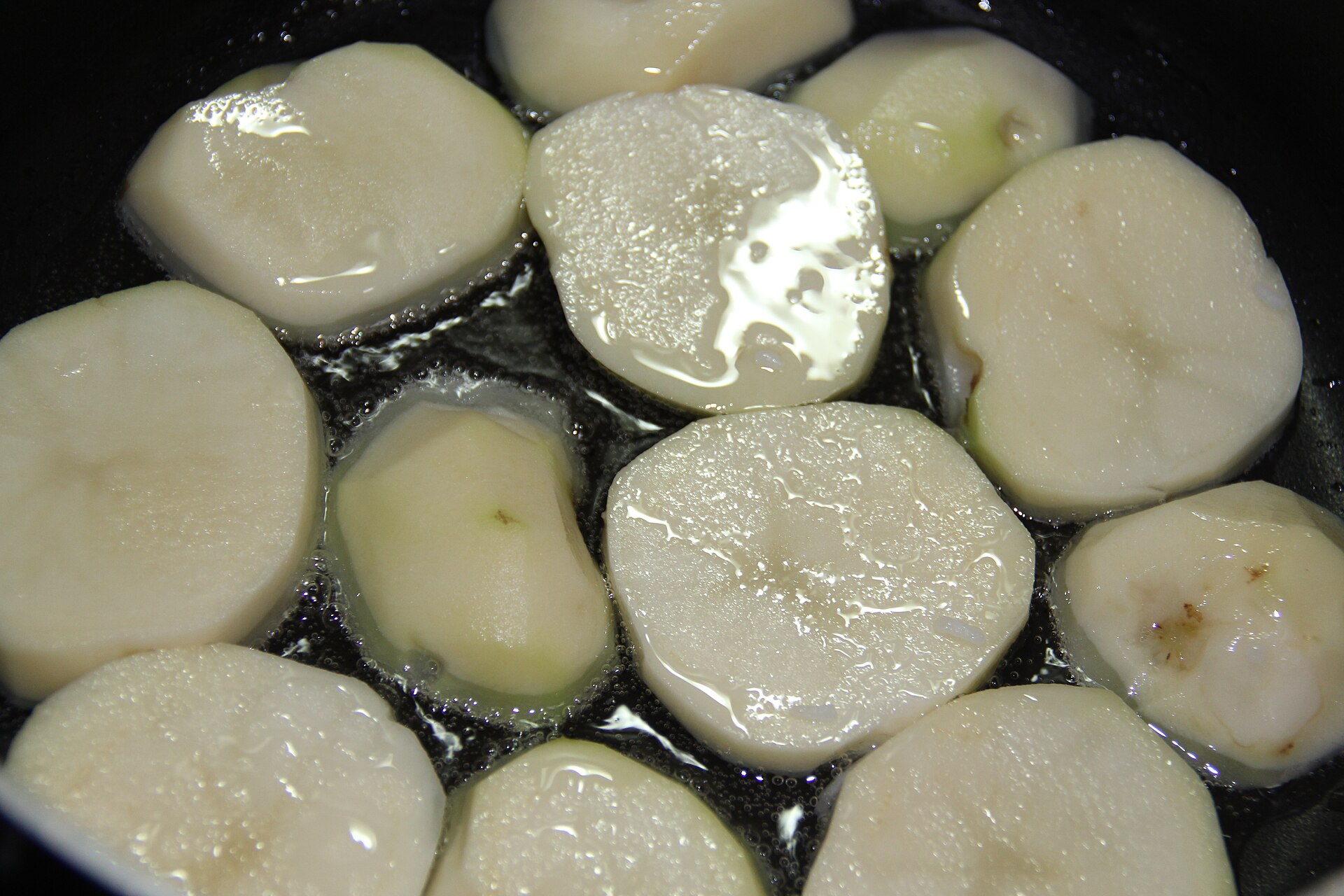 A single layer of peeled potato rounds sitting in hot oil in a dark pot.