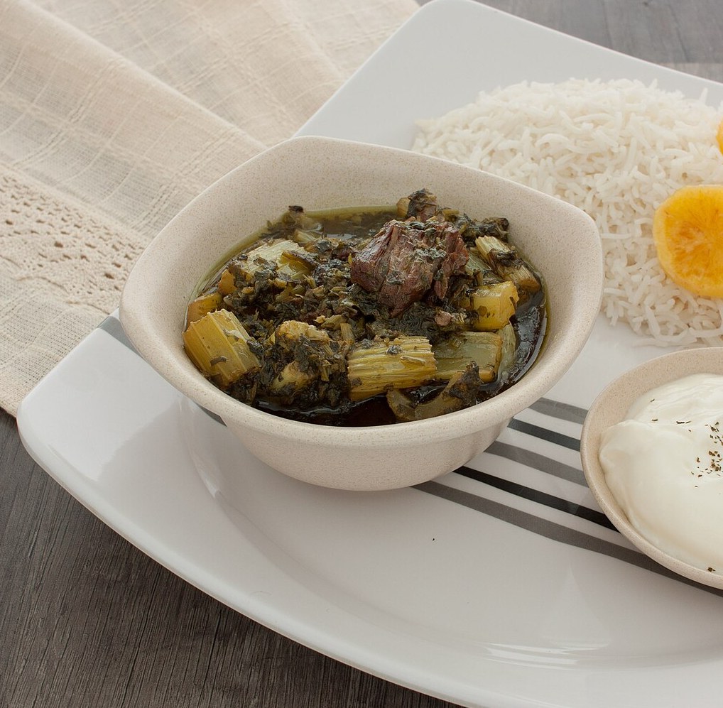 Khoresht karafs — pale green celery pieces and dark cooked herbs with lamb in a shallow bowl, served beside plain rice.