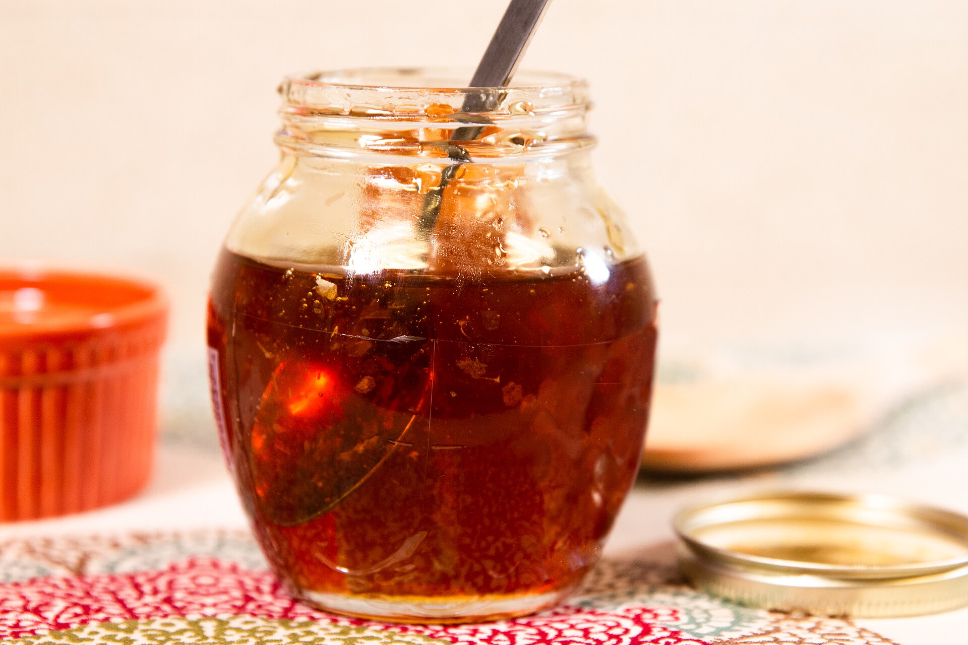 An open glass jar of amber honey with a spoon standing in it, a piece of honeycomb visible inside and the lid resting beside it