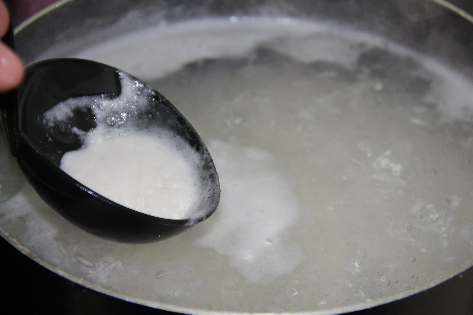 Rice boiling hard in a wide pot, foam lifted on a black spoon.
