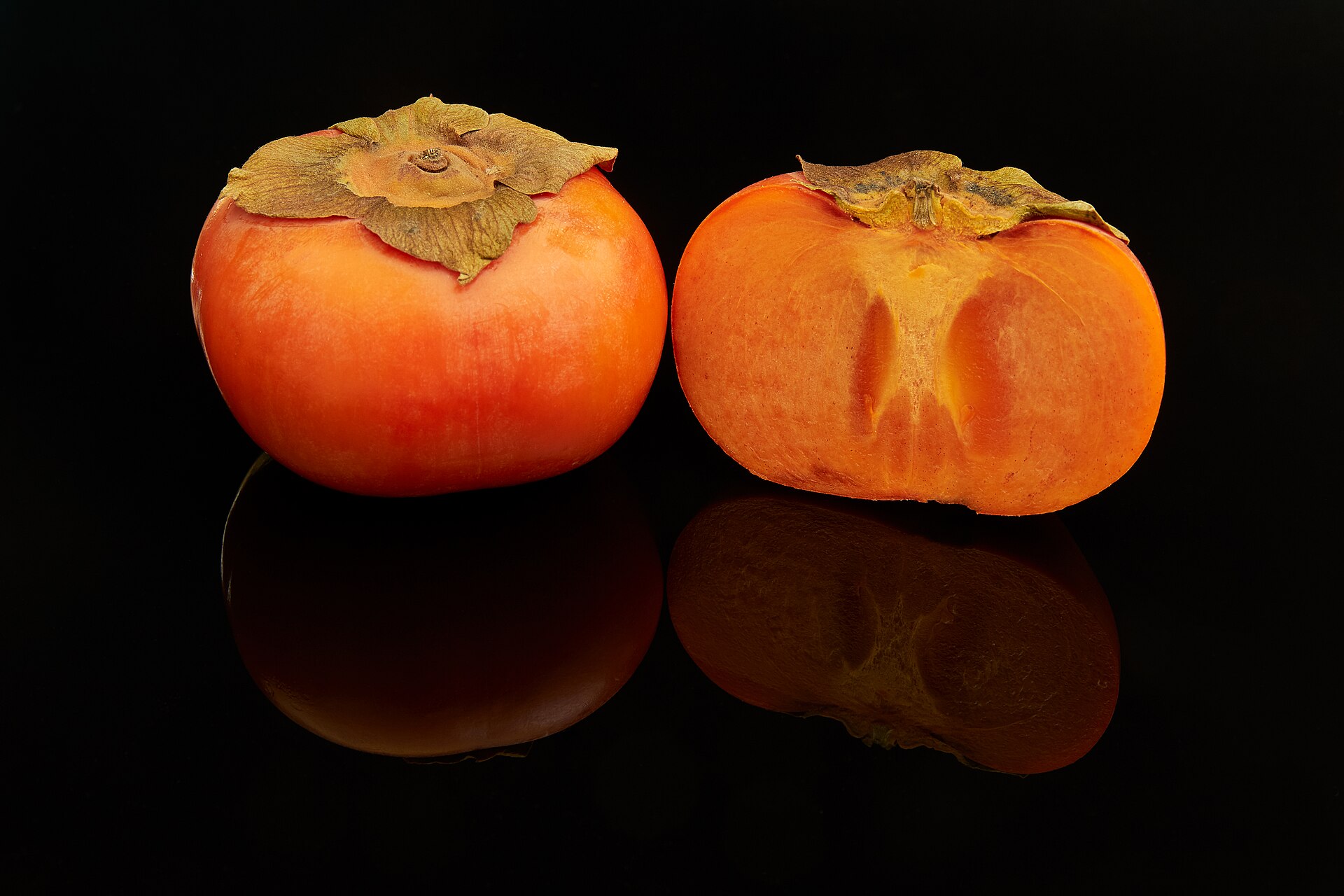 Two whole orange persimmons side by side on a dark table, one cut open to show firm pale-orange flesh