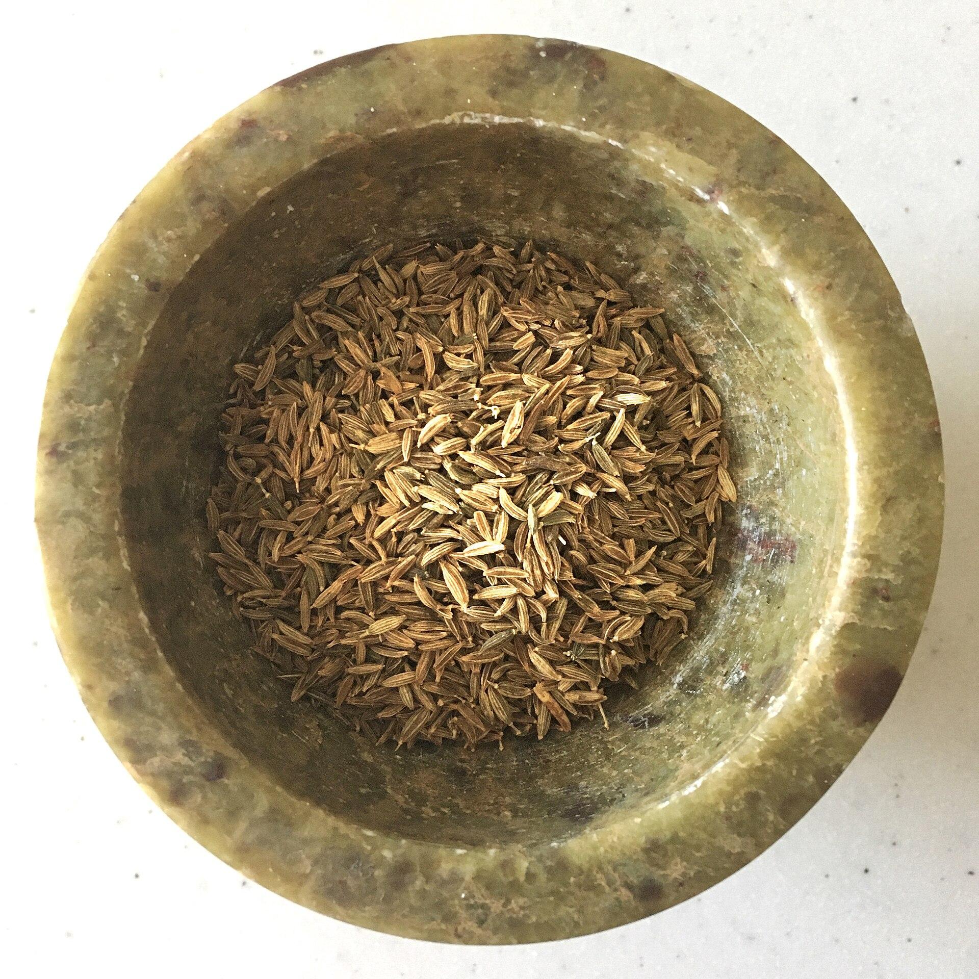 A brass mortar holding a small pile of pale ridged cumin seeds, the pestle resting beside it