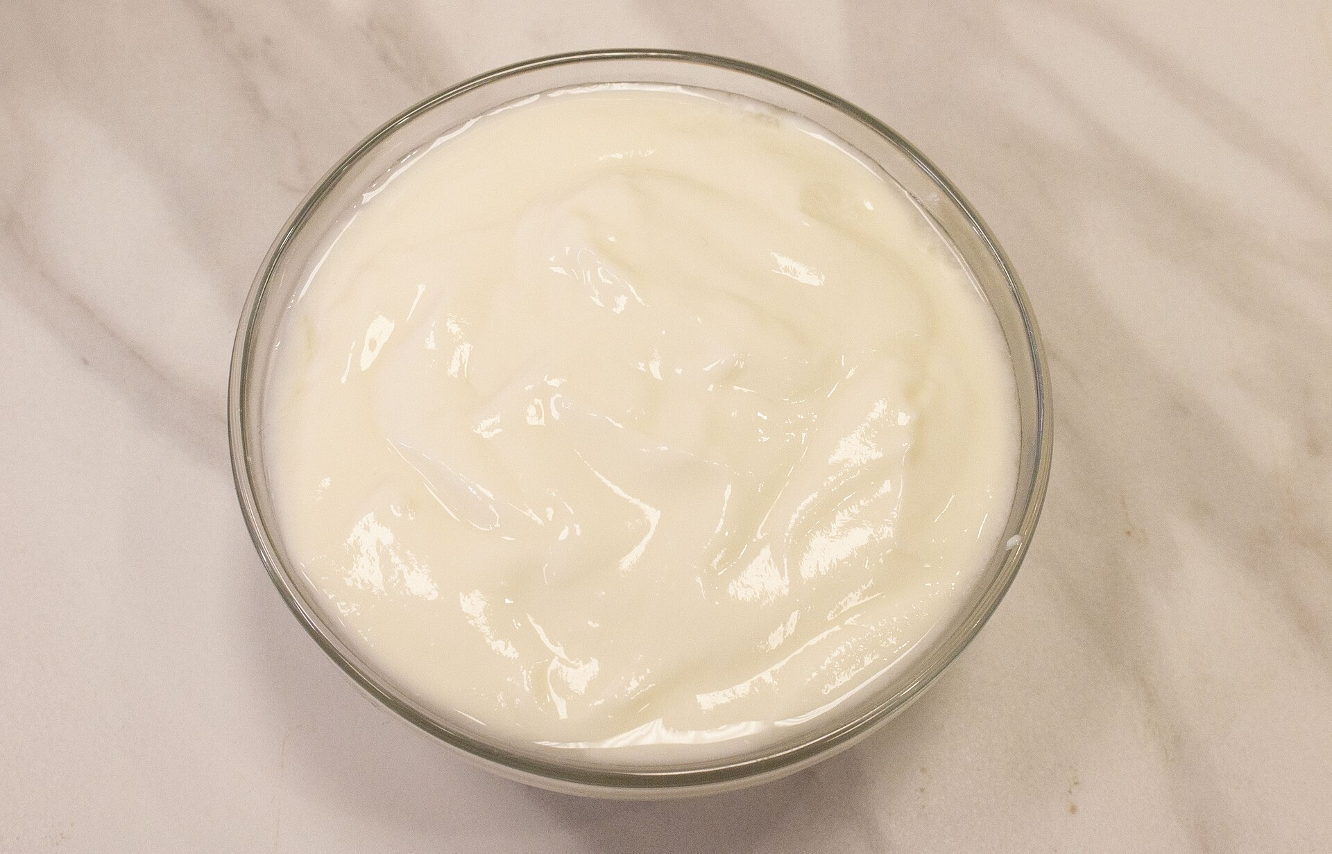 A glass bowl of thick plain white yogurt on a marble table, its surface smooth and glossy