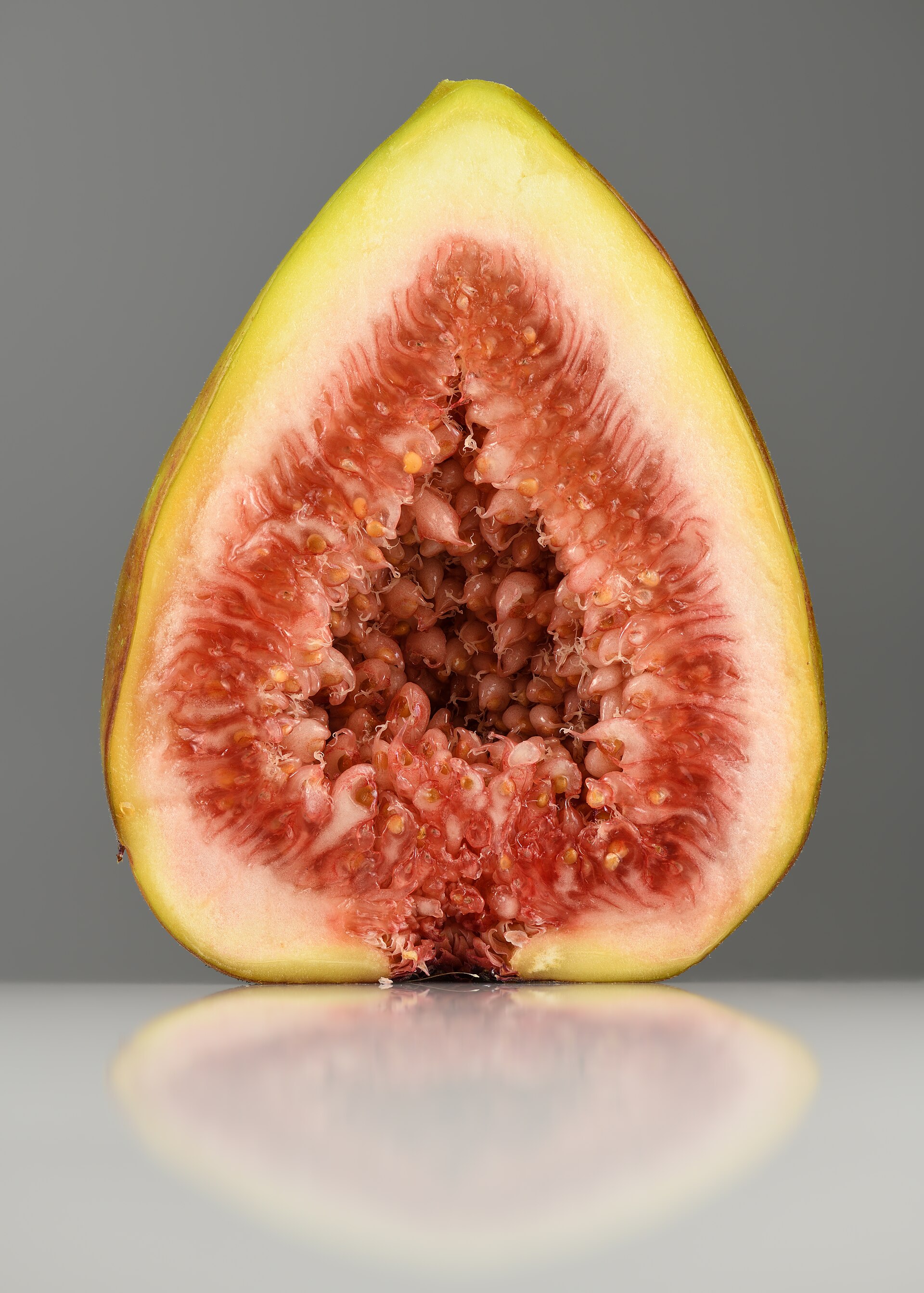 A whole fresh fig cut in half on a pale table, showing green-gold skin and dense pink seeded flesh
